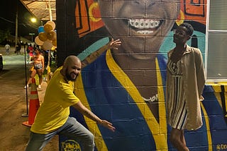 A Black man in a yellow shirt and jeans holds his hand open against a colorful mural of a woman with braces, beside a Black woman in a dress and long tan jacket also standing against a mural, which resembles her.