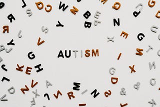 The word “AUTISM” is spelled out in the center of the image using textured, uppercase letters. The letters are colored black, gray, and brown, with the “S” and “M” in brown. Scattered around the word are numerous other individual letters in the same colors, arranged randomly on a light, neutral background.