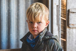 A young boy stands in front of a weathered wooden wall, wearing a camouflage jacket and a serious expression. His blond hair falls slightly over one eye, and he looks straight at the camera without smiling.