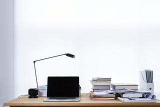 Image of laptop on a desk surrounded by piles of books and papers.