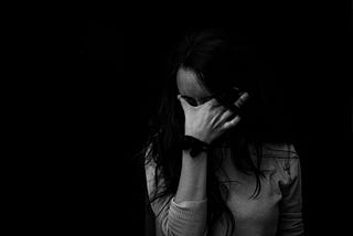 A woman stands against a black background and holds her hand against her forehead.