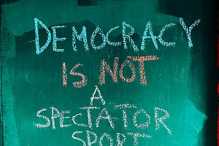 Green and black background with words in chalk writing “Democracy is not a spectator sport” — preventing democratic backsliding