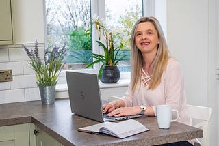 Hayley Kellard, a white female with blonde hair, sitting at a laptop, looking at the camera with a smile on her face.