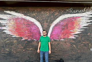 Mu’nis is a green shirt and blue jeans, standing in front of wall art depicts red and orange angel wings.