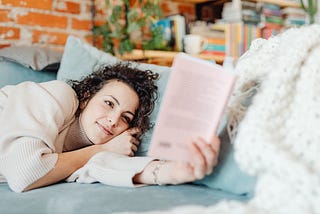 woman relaxing in bed & reading a book, with bookshelves in the background