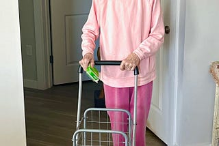 An elderly woman dressed in pink, with a pink headband, pushing a shopping cart