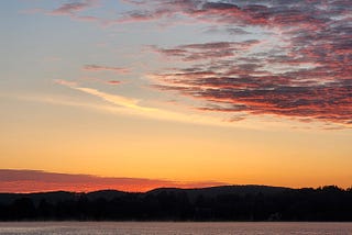 photograph of sunrise, with most of the image being the sky and clouds, though a bit of the horizon and the lake beneath is visible, with wispy fog in the distance just over the water