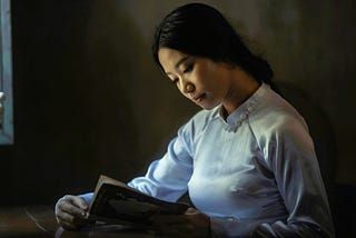 Sepia tinged photo of a young Asian woman in a high-necked white silk blouse reading a book open before her.