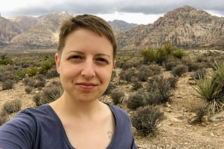Selfie photograph of a white woman with short brown hair standing in the desert. She is slightly smiling at the camera. There are rock formations in the distance and shrubby plants in the foreground.