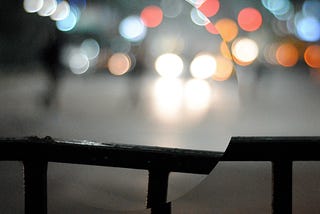 Hand rail with blurry street behind it at night with lots of lights. Part of the image is contained within a circle and rotated slightly to break the line of the hand rail.