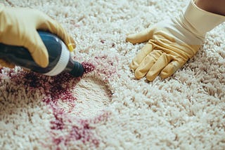 A close-up view of hands in gloves applying cleaning solution to a red wine stain on a carpet.