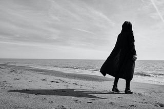 Greyscale image. Person in a black cloak stands alone on a beach near the edge of the water looking out, their shadow extending behind them.