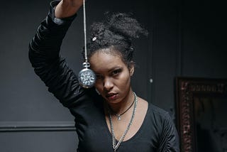 Young woman with an intense stare, dressed in black, dangling a blurry watch on a long chain in front of her face