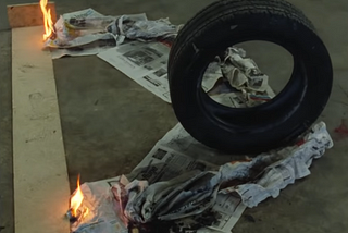 A tire sits on the tip of a v-shaped pile of newspaper. 2 flames on either side are just beginning to ignite the newspaper.