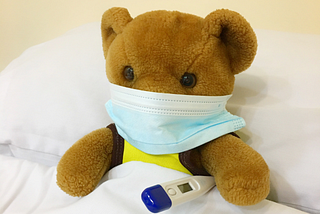 Bamse i hospitalseng med termometer og munnbind.