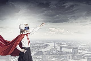 Blonde girl with a red cape and a blue mask and a white flower in her hair, white shirt, and black skirt, standing confidently with one arm outstretched Superman-style in front of a black and white city backdrop.