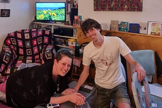 A photo of the author leaning over a padded footrest covered with a waterproof sheeting and wearing a TENS machine. They are smiling and their partner is holding their arm and smiling too. There are sunflowers on the TV behind them and other birth detritus is scattered around the room.