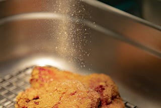 a piece of fried chicken getting peppered in a basket.