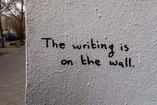 Muro branco com texto em inglês escrito “The writing is on the wall”