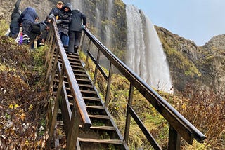Outdoors photo of tourists coming down a steep wooden stairway at the side of an Icelandic mountain with a very tall waterfall roaring in the background.