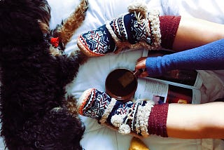 A girl’s legs wearing long socks and soft booties shown on a shite surface with a dog, a cup of tea, and a book.