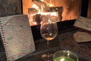 A spiral notebook sits beside stem glass of white wine and green lit candle in front of a burning wood fireplace.