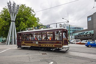 Old Invercargill Tramway tram at intersection. Coloured brown with yellow trim and modern city buildings in the background.