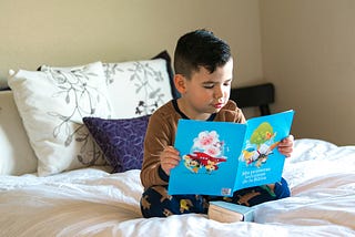 A young boy sits cross-legged on a neatly made bed, reading a bright blue children’s book. He wears brown pajamas with dark blue pants patterned with small animals. Behind him are white pillows with leafy designs and a purple accent pillow.