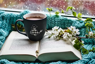 Open book with a mug of hot chocolate and white flowers over it, sitting on a blue blanket by a window on a rainy day