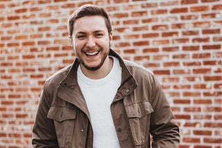 A young man smiling, in front of a brick wall.