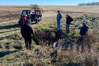 Six people stand near the edge of a field and circled around an outlet pipe.