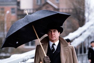A man (Alan Bates as Guy Burgess) walks under an umbrella.