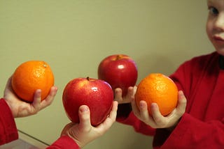 A kid holding an apple and an orange on each hand, standing in front of a mirror