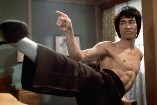 Bruce Lee dando um chute e apontando o dedo indicador na direção do adversário.