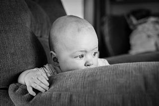 black and white. baby boy leaning over the edge of an arm chair, looking off into the distance.