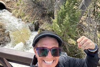 CC dressed in black with sunglasses and a black hat doing a strong arm selfie in front of a spring waterfall.