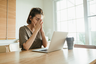 Image of woman sitting at a table in front of a computer with her hands on her face and eyes closed, apparently frustrated or worn out.