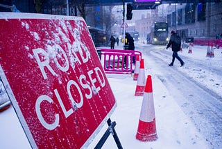 A sign viewed from its side says “Road Closed” in snowy weather next to traffic cones, a frozen road, pedestrians walking by, and a bus arriving.