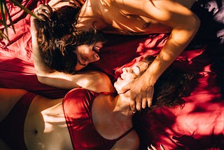 man and woman in bed with red lingerie and red sheets, smiling & in love.