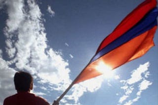 someone holds an Armenian flag against a sky with clouds