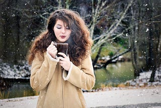 Girl, long brunette hair, winter coat, background woods and a river, blows steam off a cup of coffee