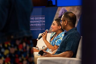A woman with short brown hair wearing a blue and yellow sundress holds a microphone while sitting in white chairs next to a man with short hair and a blue shirt on a stage at a conference.