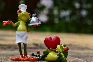 Ceramic frog chef holding a covered tray beside a frog lying at her feet holding a big red heart.