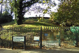 A view of the south-west corner of Claremont Square reservoir. A grassy mound and several mature trees can be seen rising behind cast-iron railings.