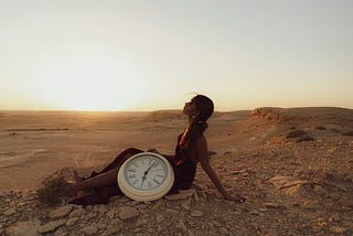 A woman is sat in a desert at sunrise with a large clock next to her