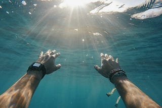 Two underwater hands reaching toward the surface and sunlight, symbolising emergence, clarity, and personal transformation.