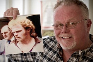 Now sporting white hair, a beard and moustache, and a receding hairline, Kirk Bloodsworth is shown holding up an older photograph of himself from the time of the initial trial. In the old photo, he has curly, reddish hair and bushy eyebrows.