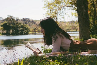 Woman reading book and lying forward on sheet on grass beside body of water during the day.E.g.