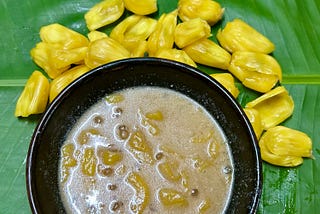 A black bowl with brownish yellow liquid featuring granules is placed over a banana leaf, yellow coloured jackfruit bulbs surround the bowl.