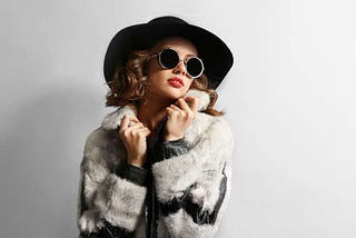 Young woman wearing a faux-fur coat, hat, and sunglasses, to replicate the mob-wife aesthetic